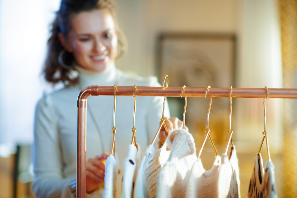 Closeup on stylish woman in white sweater and skirt at modern home in sunny winter day choosing sweaters hanging on copper clothes rack.